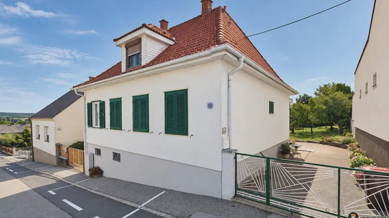 Traditionelles Wohnhaus mit weißer Fassade, roten Dachziegeln, grünen Fensterläden und Gartenbereich.