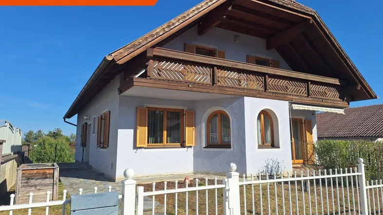 Traditionelles Einfamilienhaus mit Holzbalkon und gepflegtem Vorgarten unter blauem Himmel.