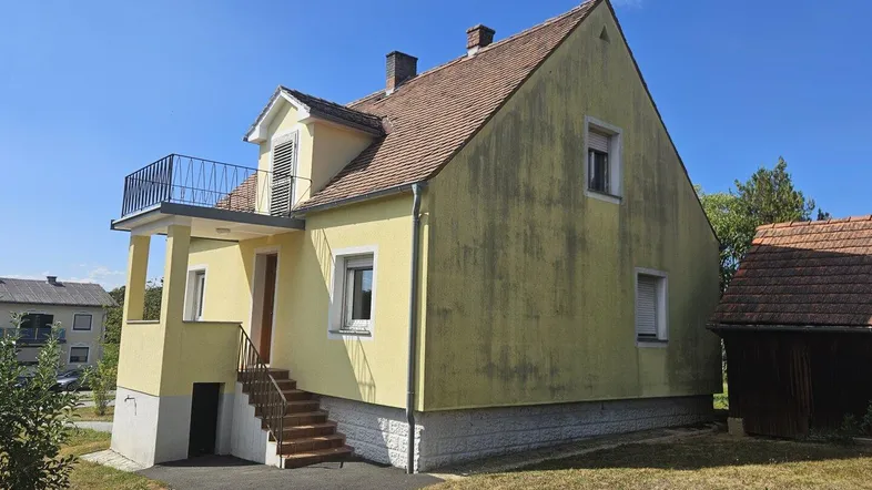 Gelbes Einfamilienhaus mit Balkon, Satteldach und Außentreppe zum Kellerzugang in ländlicher Umgebung.