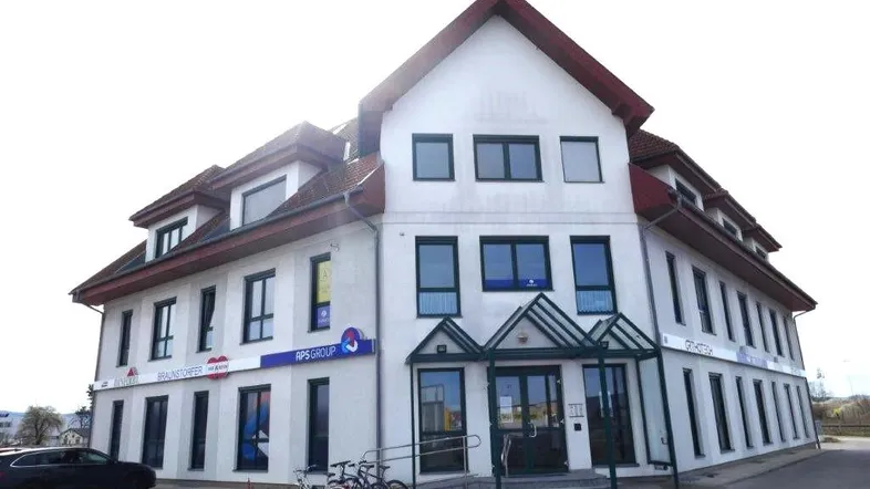 Gebäudeansicht mit Haupteingang, Rampe und mehreren Fenstern unter einem roten Ziegeldach.