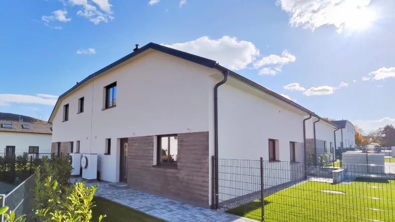 Moderne Doppelhaushälfte mit heller Fassade, dunklem Dach und gepflegtem Rasen unter blauem Himmel.