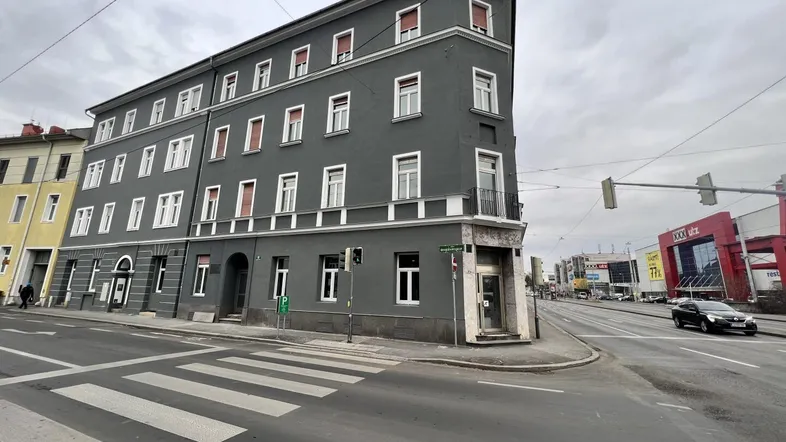 Graues Eckgebäude an einer belebten Straße mit vielen Fenstern und einem Zebrastreifen im Vordergrund.