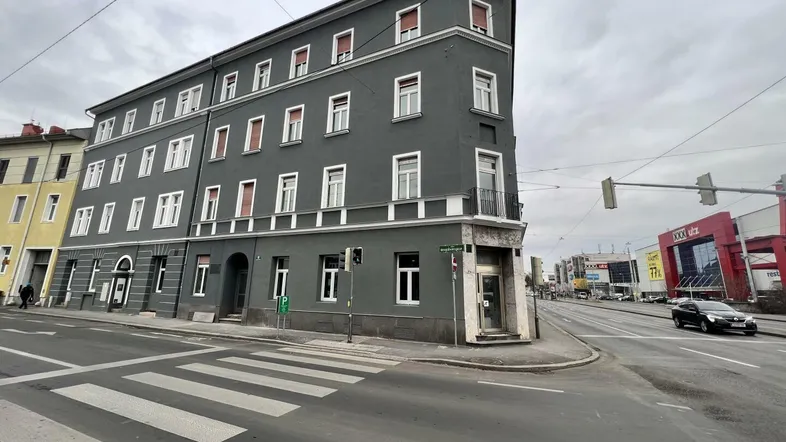 Das Gebäude präsentiert sich als Eckhaus mit einer dunkelgrauen Fassade und vielen Fenstern an einer städtischen Kreuzung.