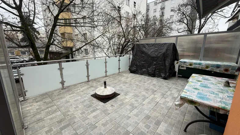 Geräumige Terrasse mit Fliesenboden und Glasgeländer, umgeben von städtischer Bebauung und Bäumen.