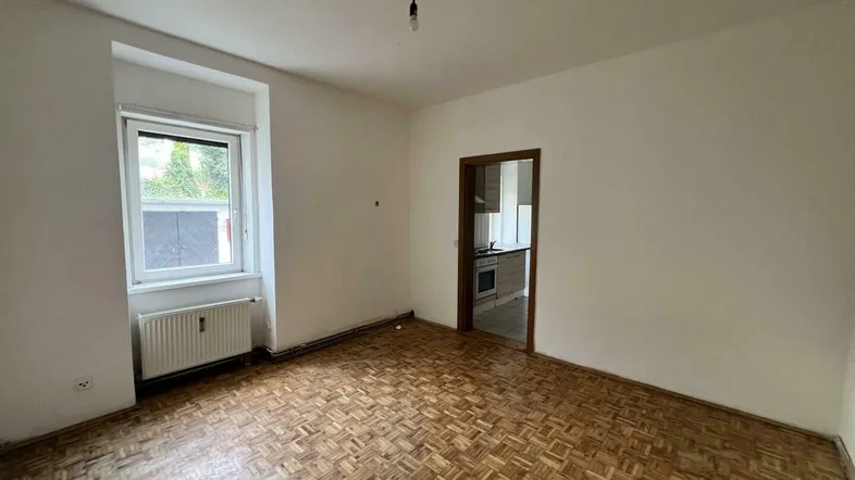 Helles Zimmer mit Fischgrätparkettboden und Fenster, das als Wohn- oder Schlafbereich genutzt werden kann.