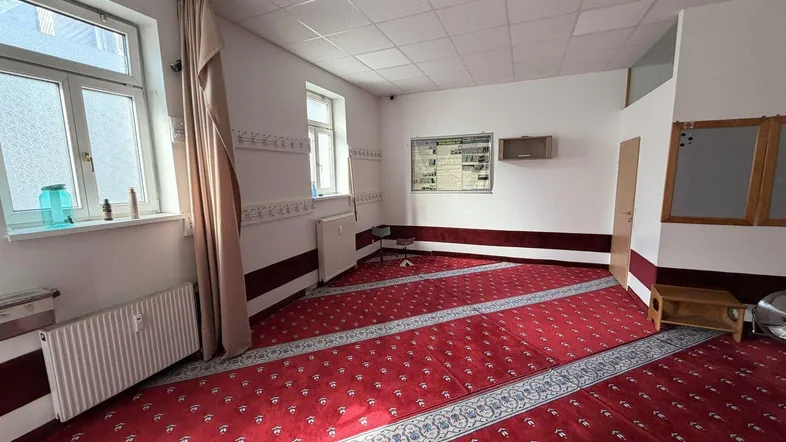 Geräumiger Innenraum mit rotem Teppichboden, weißen Wänden und mehreren Fenstern, ideal für Versammlungen.