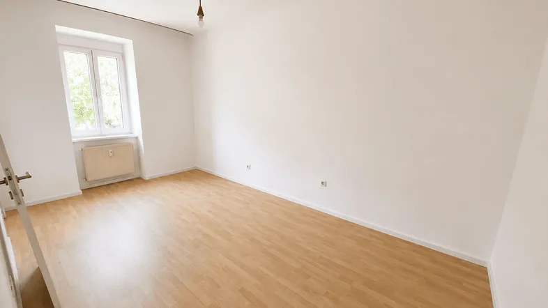 Helles, leeres Zimmer mit Laminatboden und einem Fenster, das viel Tageslicht hereinlässt.
