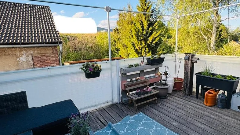 Sonnige Holzterrasse mit Outdoor-Sofa, Pflanzen und weitem Blick ins Grüne.