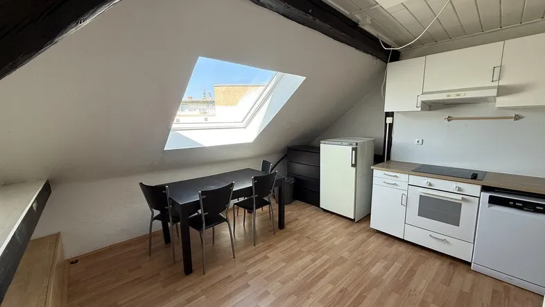 Helle Dachgeschosswohnung mit kleiner Einbauküche, Essbereich und sichtbaren Holzbalken unter der Dachschräge.