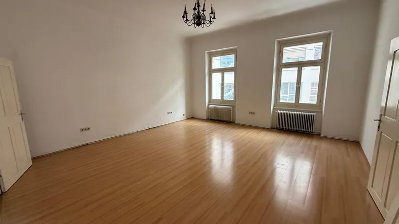 Geräumiges, leeres Zimmer mit hellem Holzboden, weißen Wänden und zwei großen Fenstern.