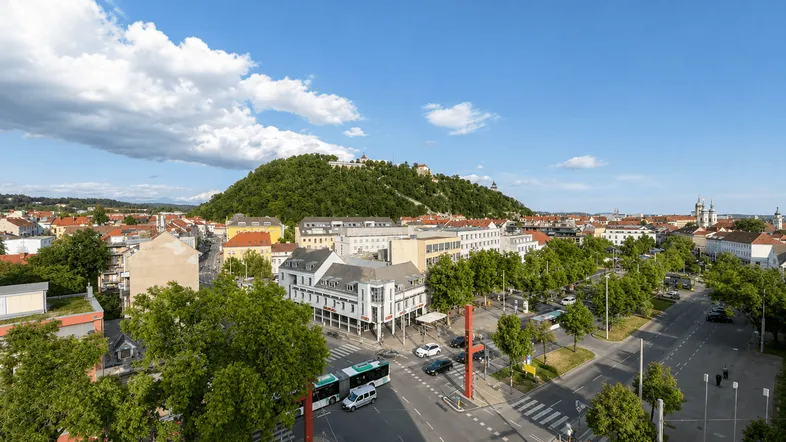 Panoramablick über die Stadt mit grünen Hügeln, Gebäuden und belebten Straßen unter blauem Himmel.