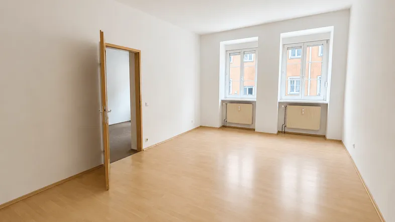 Helles Wohnzimmer mit Laminatboden, hohen Decken und zwei Fenstern, die viel Tageslicht hereinlassen.