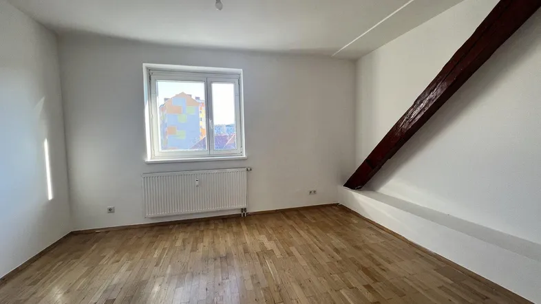 Helles Zimmer im Dachgeschoss mit Holzbalken, Parkettboden und einem Fenster mit Blick nach draußen.
