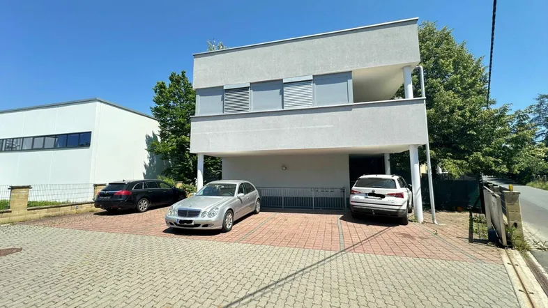 Modernes Gebäude mit Flachdach und Balkonen, davor ein gepflasterter Parkplatz mit mehreren Autos.