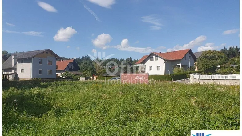 Ungenutztes Grundstück mit grüner Wiese und Wohnhäusern im Hintergrund unter blauem Himmel.