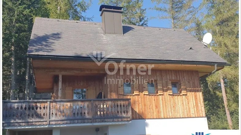 Traditionelles Holzhaus mit Satteldach und einem charmanten Balkon, umgeben von hohen Nadelbäumen.
