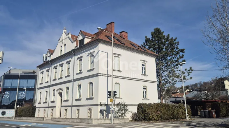 Weißes Altbaugebäude mit rotem Ziegeldach an einer Straßenecke unter blauem Himmel.