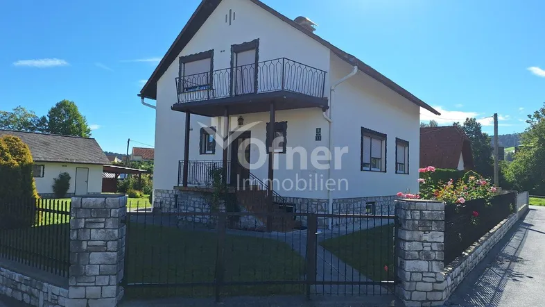 Vorderansicht eines Einfamilienhauses mit zwei Balkonen, umzäuntem Vorgarten und sonnigem Wetter.