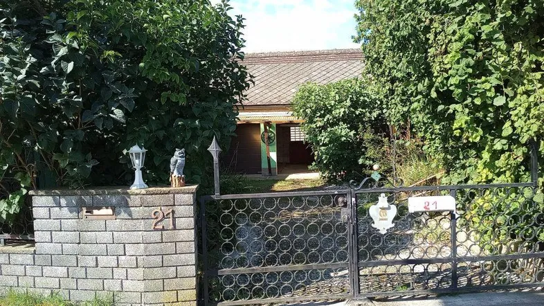 Vorderansicht eines Hauses hinter einem Zaun und üppigem Grün, mit einer Einfahrt.