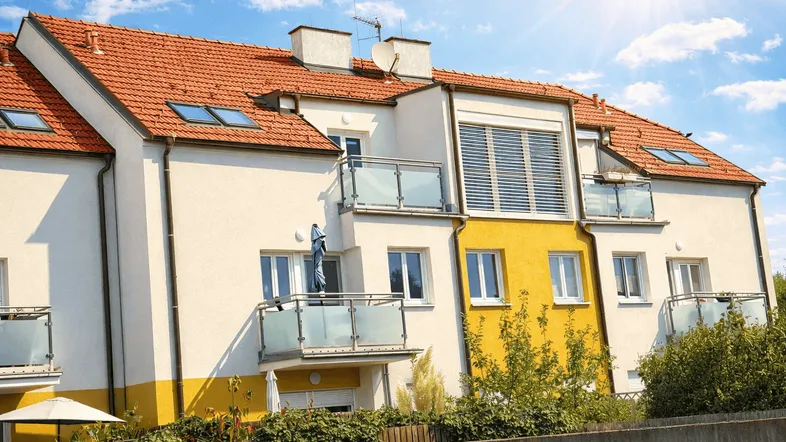 Mehrfamilienhaus mit gelber Fassade, roten Ziegeldächern und mehreren Balkonen unter strahlendem Himmel.