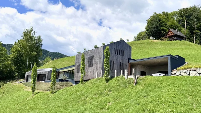 Modernes Haus mit Holzfassade und begrüntem Dach, eingebettet in eine hügelige Landschaft.