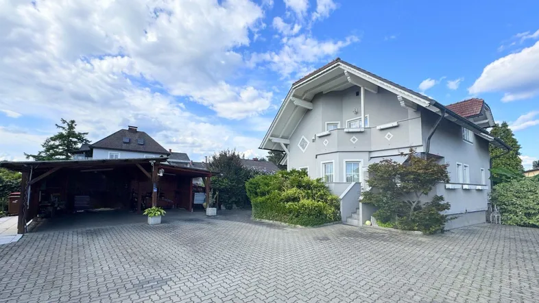 Graues Einfamilienhaus mit gepflasterter Auffahrt und überdachtem Carport unter blauem Himmel.