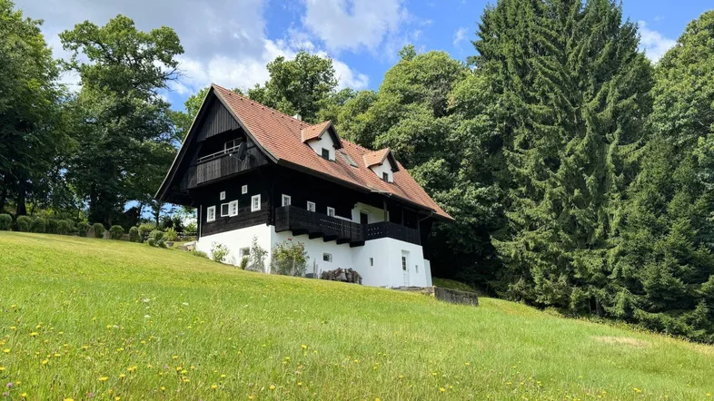 Traditionelles Haus mit Holzbalkonen und weißer Fassade, umgeben von grünen Bäumen und Wiesen.