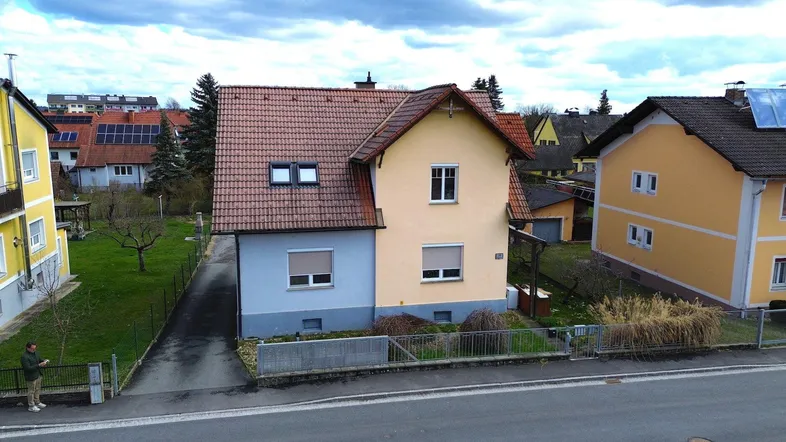 Ansicht des Hauses von der Straße mit Vorgarten und angrenzenden Gebäuden unter bewölktem Himmel.