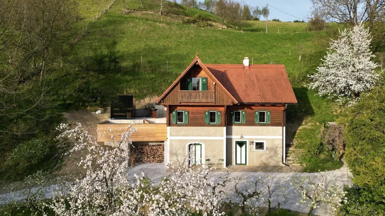 Haus in ländlicher Umgebung mit blühenden Bäumen und grünen Hügeln im Hintergrund.
