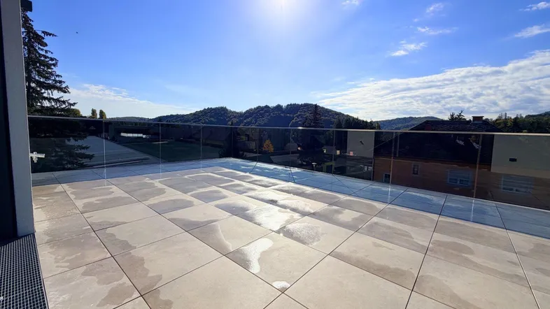 Große, sonnige Terrasse mit Fliesenboden und Glasgeländer, bietet einen weiten Blick auf die umliegende Landschaft.