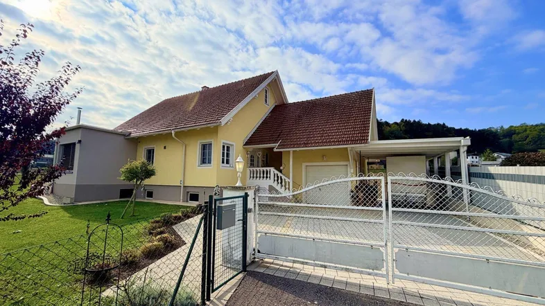 Gelbes Einfamilienhaus mit Satteldach, Vorgarten und Einfahrt unter blauem Himmel.