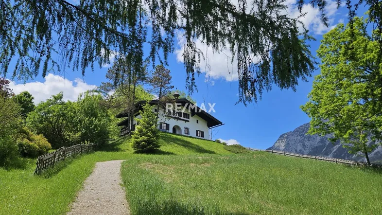 Charmantes Haus auf einem grünen Hügel mit Bäumen und Bergen im Hintergrund unter blauem Himmel.
