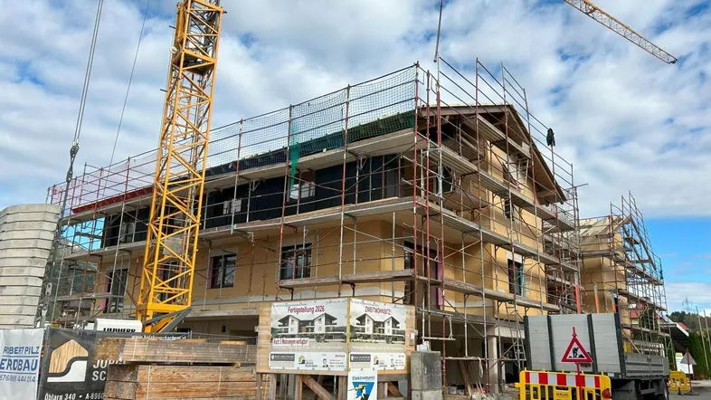 Neubauprojekt im Bau mit Gerüst, Kran und Bauschildern vor bergiger Kulisse.
