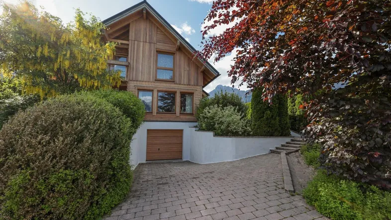 Modernes Holzhaus mit integrierter Garage und gepflasterter Auffahrt, umgeben von üppigem Grün.