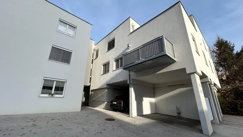 Modernes Mehrfamilienhaus mit weißer Fassade, Balkon und überdachtem Parkplatzbereich.