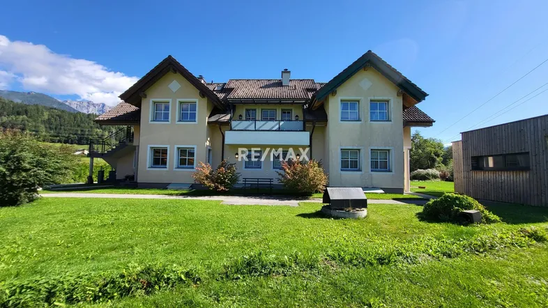 Großzügiges Haus mit gepflegtem Vorgarten und Bergblick unter blauem Himmel.