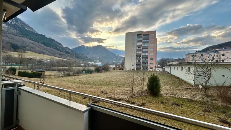 Blick vom Balkon auf eine grüne Wiese mit Bergen im Hintergrund und einem Wohngebäude.