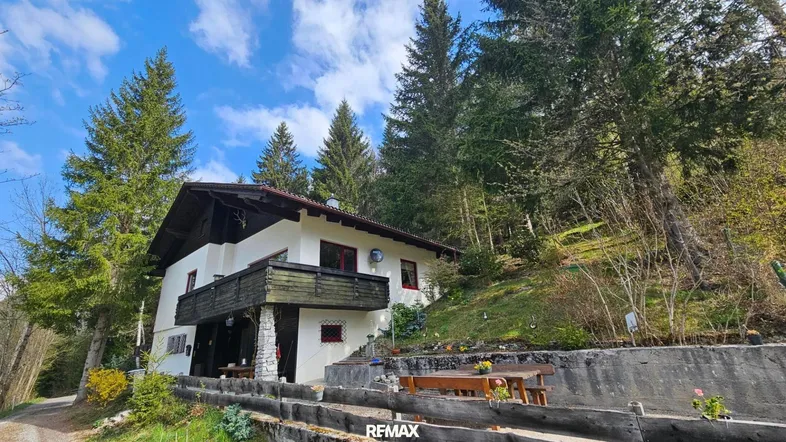 Traditionelles Haus im alpinen Stil mit Balkon, umgeben von grünen Bäumen und einem sonnigen Garten.