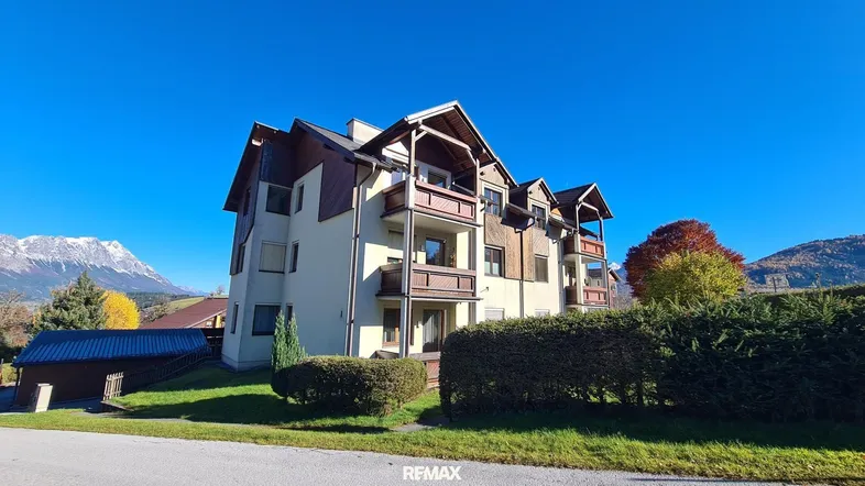 Mehrfamilienhaus mit traditioneller Architektur, Balkonen und Bergblick unter blauem Himmel.
