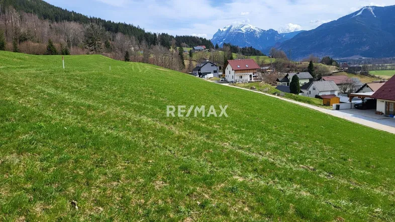 Weitläufige grüne Wiese mit Blick auf umliegende Häuser und schneebedeckte Berge im Hintergrund.