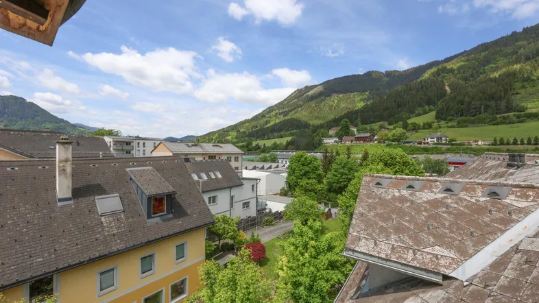 Weitläufiger Blick über Dächer und grüne Hügel, mit Bergen im Hintergrund unter blauem Himmel.