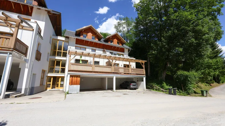 Mehrstöckiges Gebäude mit Holzfassade, Balkonen und überdachten Parkplätzen in grüner Umgebung.