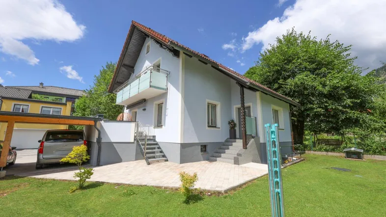 Freistehendes Haus mit hellblauer Fassade, gepflegtem Garten und Carport unter blauem Himmel.