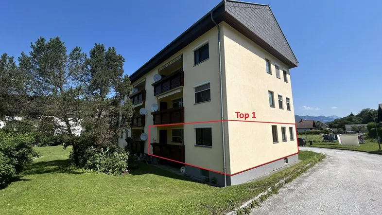 Mehrfamilienhaus mit gelber Fassade und Balkonen in ländlicher Umgebung. Die Wohnung "Top 1" ist markiert.