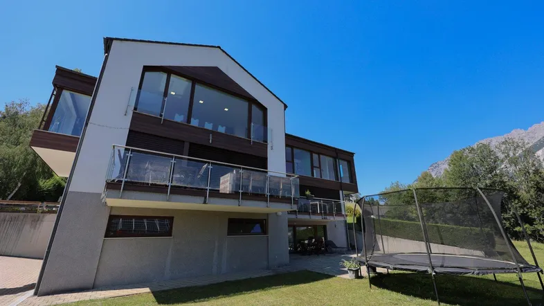 Gartenbereich mit Trampolin und moderner Hausfassade vor Bergkulisse.