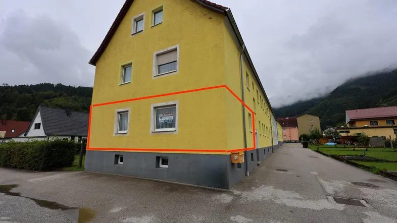 Gelbes Mehrfamilienhaus mit roter Umrandung und Bergblick unter bewölktem Himmel.