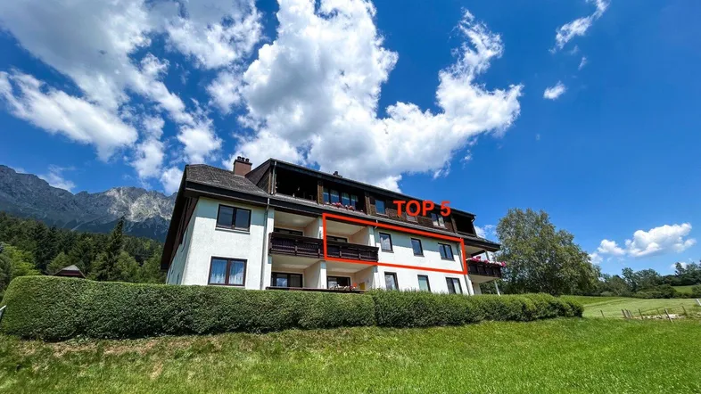Mehrfamilienhaus in ländlicher Umgebung mit Balkonen und Bergblick unter blauem Himmel.