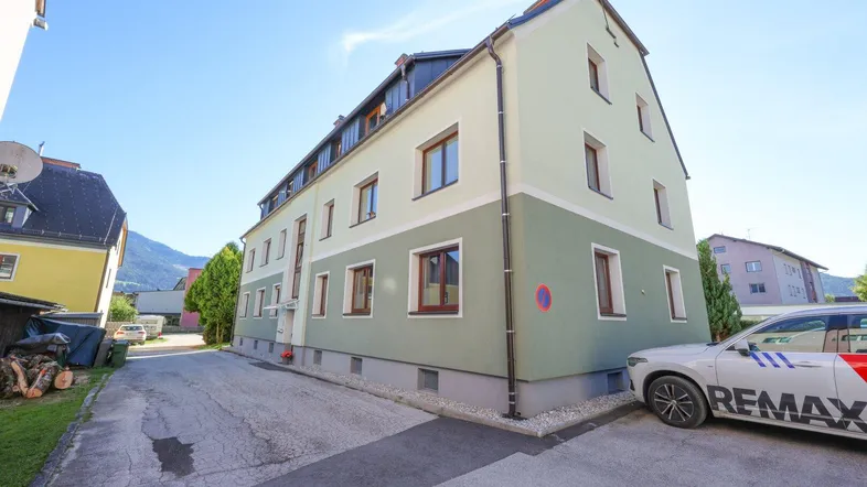 Gebäudeansicht mit Parkplatz und grüner Fassade unter blauem Himmel an einem sonnigen Tag.