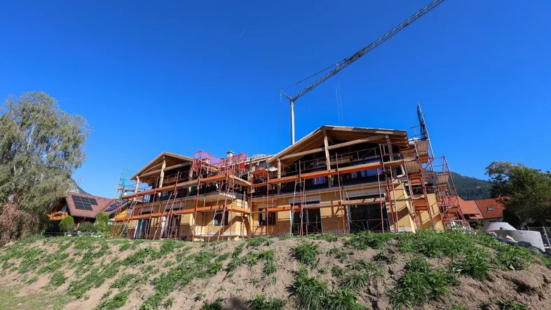Neubau im Rohbau mit Gerüst und Kran vor blauem Himmel, umgeben von grüner Landschaft.