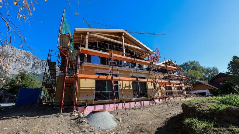 Neubau im Rohbau mit Gerüst und sichtbarer Holzkonstruktion vor Bergkulisse bei strahlendem Sonnenschein.